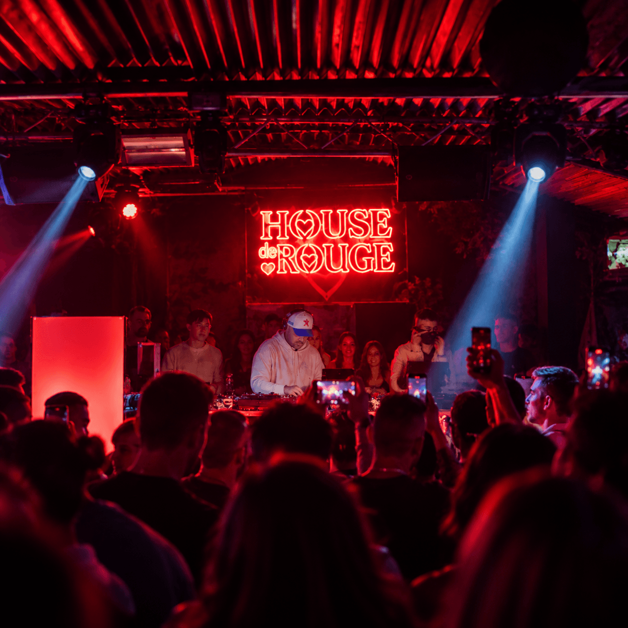 DJ mit weißer Cap am Pult unter der roten Neonschrift „House de Rouge“, Scheinwerferkegel über der Menge mit erhobenen Handys