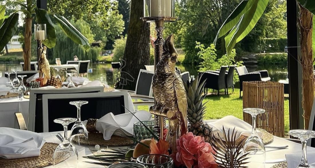 Gedeckte Gartentafel am Wasser: goldene Papageienfigur und Kerzenleuchter zwischen Ananas, Blüten und Palmwedeln, dahinter die Terrasse und der Fluss