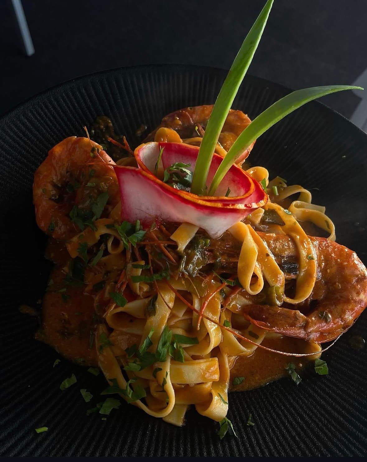 Tagliatelle mit Scampi in Safran-Tomatensugo auf schwarzem Rillenteller, garniert mit einer Radieschenblüte und Lauchspitzen